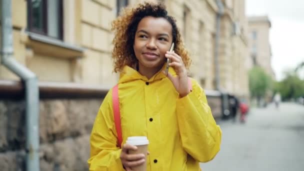 Cheerful mixed-race teenage girl is talking on smart phone and holding take out coffee walking along street in beautiful city. Modern lifestyle and technology concept. — Stock Video