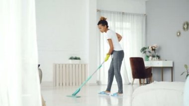 Genç Afrikalı-Amerikalı kadın paspas ile modern ev zemin temizleme, ev giyim, terlik ve lastik eldiven giymiş güzel kız çalışmalarına konsantre.