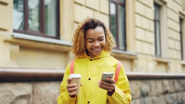 Atractiva mujer de raza mixta está utilizando un teléfono inteligente moderno mirando a la pantalla y sonriendo navegar por la red o ver fotos y la celebración de tomar café de pie al aire libre . — Vídeos de Stock