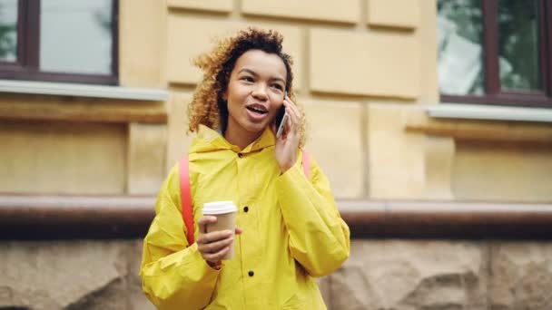 Une Afro-Américaine négligente parle sur son portable et tient un café à emporter debout à l'extérieur dans une ville moderne. Concept de communication et technologie . — Video