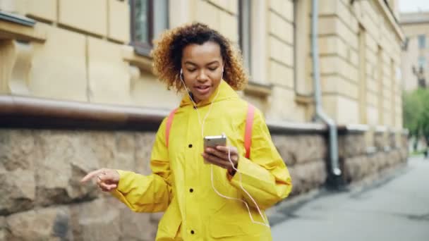 若いアフリカ系アメリカ人女性はイヤホンを介して音楽を聴くと踊り明るい色の服を身に着けている現代の都市の通りに沿って歩きます。楽しさとガジェットのコンセプト. — ストック動画