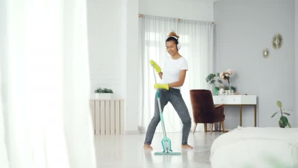 Divertido adolescente afroamericano está limpiando la casa haciendo tareas domésticas limpiando el piso con fregona y escuchando música en los auriculares, chica está bailando y cantando . — Vídeo de stock