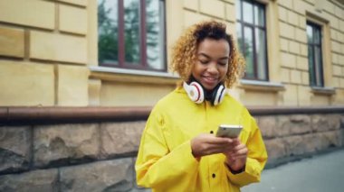 Mutlu Afrika kökenli Amerikalı öğrenci smartphone tarama veya manifatura arkadaşlar açık havada kulaklıklar giyen ve gülümseyen modern şehir sokakta duran kullanıyor.