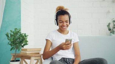 Mutlu Afrikalı-Amerikalı kadın smartphone ve evde koltukta oturan gülüyor ile internet sörf, kız kulaklık aracılığıyla müzik dinlerken.