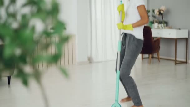 Divertida chica afroamericana está disfrutando de la música en los auriculares y haciendo tareas domésticas lavando piso en casa, joven dama está bailando y cantando divertirse . — Vídeo de stock