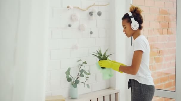 Vrolijke meid African American meisje doet huishoudelijk werk afstoffen met doek en het dragen van handschoenen, jonge vrouw is luisteren naar muziek in hoofdtelefoons, dansen en zingen. — Stockvideo