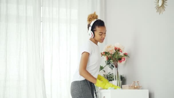 La señora de la limpieza afroamericana en guantes protectores está espolvoreando los muebles con tela húmeda y escuchando música con auriculares y luego bailando y cantando . — Vídeo de stock