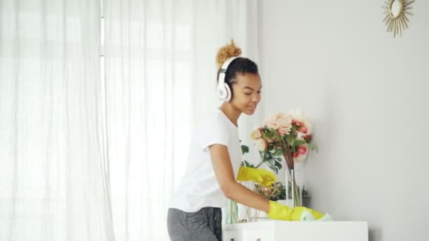 Ziemlich gemischte Rasse Hausfrau staubt die Möbel und genießt Musik mit Kopfhörern, tanzen und singen. glückliche junge Frau hat Spaß und macht Hausarbeit. — Stockvideo