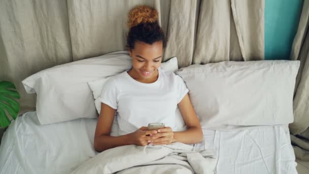 Senhora afro-americana alegre usando smartphone deitado na cama sob cobertor quente em travesseiro macio. Conceito de hora de dormir, comunicação e tecnologia moderna . — Vídeo de Stock