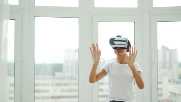 Chica afroamericana en ropa casual está usando gafas de realidad virtual y gestos mientras está de pie en su apartamento. Tecnología moderna, entretenimiento y concepto de personas . — Vídeos de Stock
