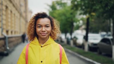 Gülümseyen ve kamera açık havada yeşil ağaçlar ve arka planda güzel binalar ayakta seyir mutlu Afro-Amerikan Kız portresi ağır çekim.