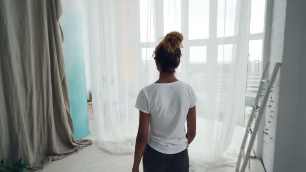 Vista trasera de la mujer afroamericana caminando hacia la ventana panorámica, separando las cortinas y mirando hacia el exterior disfrutando de la vista de la gran ciudad moderna. Alojamiento y concepto de personas . — Vídeo de stock
