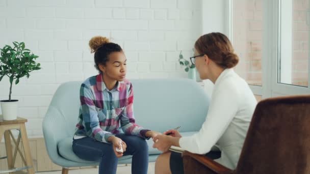 Känslomässiga African American lady pratar med psykologen sitter på soffan, prata och gestikulerande under samrådet medan specialist lyssnar hålla papper och penna. — Stockvideo