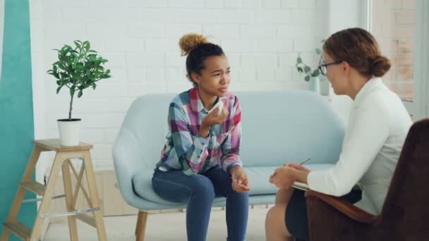 Unhappy mixed race girl is talking to psychologist and crying sitting on couch while doctor is listening to her holding paper and pen. Therapy, people and problems concept. — Stock Video