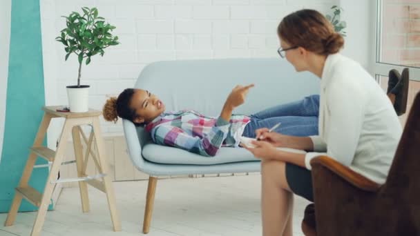 Afroamerikanska tonåringen pratar med psykologen liggande på soffan och ler medan terapeuten lyssnar och antecknar. Ungdomar, psykisk hälsa och yrke koncept. — Stockvideo