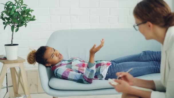 意気消沈したアフリカ系アメリカ人女の子はソファーに横になっていると、メガネのセラピストは、リスニング、ライティング、スピーキングの心理学者に話しています。ヘルプとスペシャ リストの概念. — ストック動画