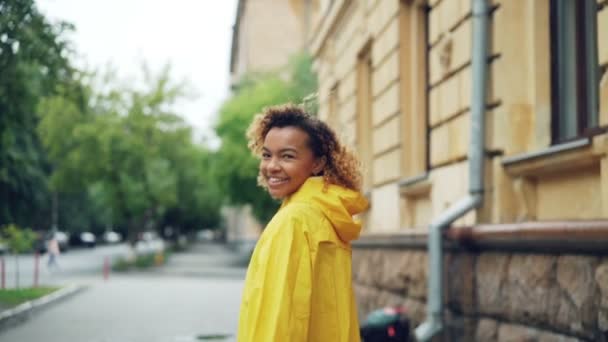 Retrato en cámara lenta de una mujer afroamericana sonriente con un abrigo brillante caminando por la calle, girando hacia la cámara y mirando a la cámara y luego girando . — Vídeos de Stock