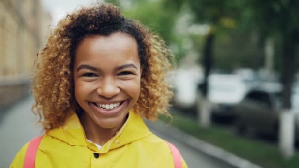 Primer plano retrato en cámara lenta de una mujer afroamericana guapa mirando a la cámara con sonrisa alegre y riendo expresando emociones positivas de pie afuera . — Vídeo de stock