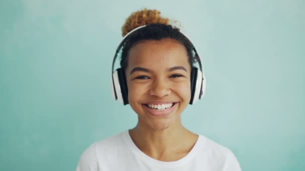 Close-up retrato em câmera lenta de menina afro-americana feliz em fones de ouvido olhando para a câmera, sorrindo e rindo. Pessoas, tecnologia moderna e conceito divertido . — Vídeo de Stock