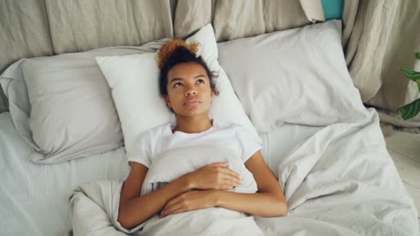Pretty mixed race lady is lying in bed and sighing looking at ceiling then closing her eyes and falling asleep with smile. Comfortable bed, pillows and sheets are visible. — Stock Video