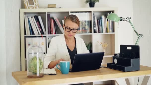 Mujer joven atractiva está trabajando en la oficina moderna sentada en la mesa, utilizando el ordenador portátil y la escritura en el cuaderno. Estantes con papeles, plantas y libros son visibles — Vídeos de Stock