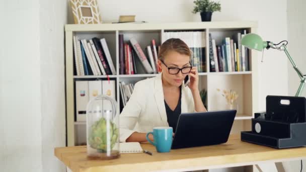Αυτοπεποίθηση γυναίκα υπάλληλο της εταιρείας είναι να μιλάμε στο κινητό τηλέφωνο, χρησιμοποιώντας το laptop και τις σημειώσεις στο σημειωματάριο που κάθεται στο γραφείο στο χώρο εργασίας. Επιχειρήσεων, γραφείων και άνθρωποι έννοια. — Αρχείο Βίντεο