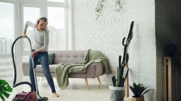 Glada blond hemmafru använder dammsugare under rutinmässiga städa hemma, lyssna på musik via hörlurar, sång och dans. Teknik och människor-konceptet. — Stockvideo