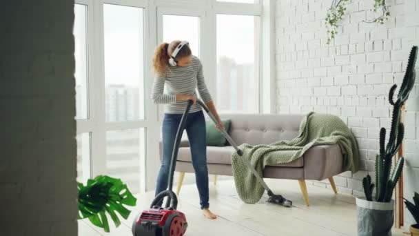 Belle femme vole le sol à la maison en utilisant un aspirateur moderne et en écoutant de la musique avec écouteurs, danse et chant. Concept de ménage et de technologie . — Video
