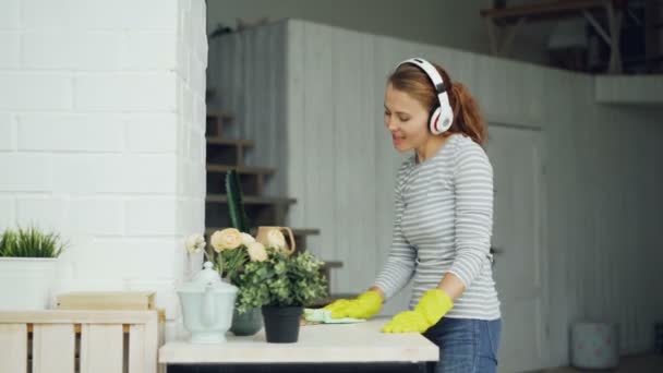 Chica guapa está limpiando la mesa en casa y escuchando música a través de auriculares, cantando y bailando con la cara feliz. Millennials, concepto de hogar e interior . — Vídeo de stock