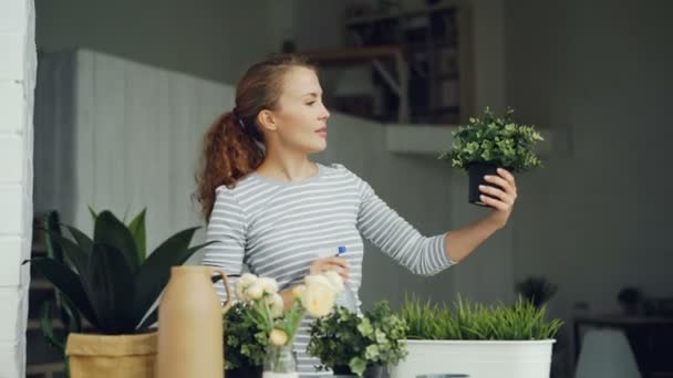 Neşeli ev kadını Çiçeklik tutarak ve güzel ışık daire ayakta gülümseyen püskürtücü kullanarak yeşil bitkilerin sulama. Ev işleri ve botanik kavramı. — Stok video