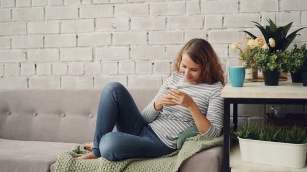 Attractive girl in casual clothing is using smartphone sitting on sofa in modern loft style apartment and smiling. Modern technology, interior and youth concept. — Stock Video