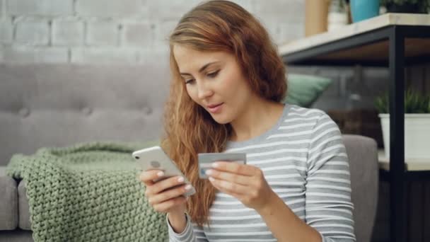 Jolie jeune femme fait du shopping en ligne en payant ses achats par carte bancaire à l'aide d'un smartphone, la fille tient la carte et entre les informations à l'écran tactile puis sourit . — Video