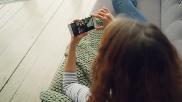 Vista trasera de la señorita skyping con amigos hermosa pareja con teléfono inteligente sentado en el sofá en casa. Tecnología moderna, millennials y concepto de comunicación . — Vídeos de Stock