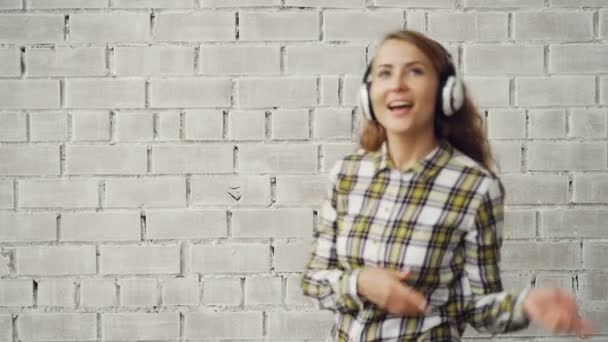 Portrét atraktivní dívka poslechu hudby přes sluchátka, zpěvem a tancem proti cihlové zdi pozadí. Koncepce mládeže milénia, kultura a životní styl. — Stock video