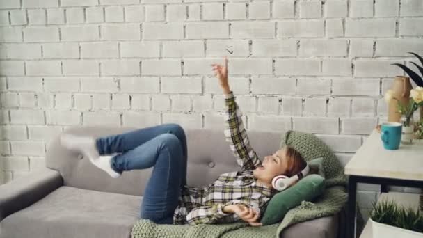 Joyful young woman is singing, listening to music in headphones and moving hands and legs dancing lying on sofa in modern loft style apartment. People and fun concept. — Stock Video