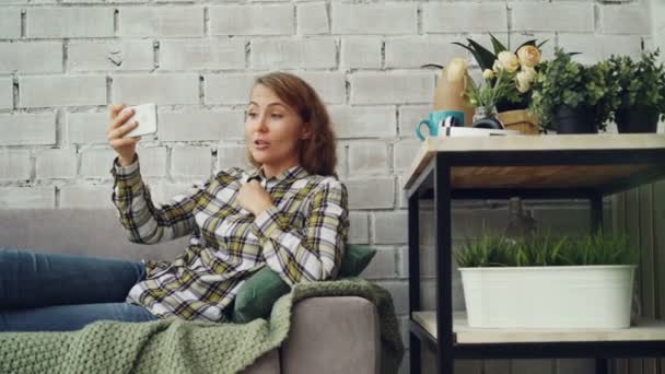 Bella donna sta chattando con gli amici guardando lo schermo dello smartphone e parlando con sorriso felice seduto sul divano in un moderno appartamento in stile loft. Tecnologia e concetto di gioventù . — Video Stock