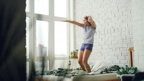Vrolijke jonge vrouw is dansen op bed thuis hoofdtelefoon dragen en genieten van muziek. Dubbel bed, bakstenen muur, panoramisch venster en groene plant zijn zichtbaar. — Stockvideo