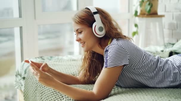 Mulher jovem bonito está ouvindo música em fones de ouvido e tocando tela do smartphone descansando na cama em casa. Tecnologia, cultura juvenil e conceito de interiores . — Vídeo de Stock