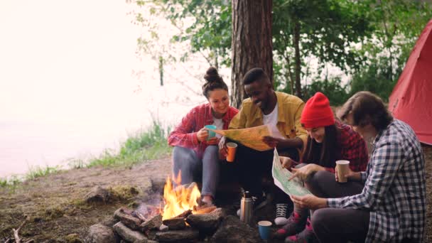 Boucle cinématographique - un groupe de jeunes hommes et de jeunes femmes regarde des cartes papier autour d'un feu dans la forêt avec des boissons. Carte en mouvement, tente et arbres sont visibles . — Video