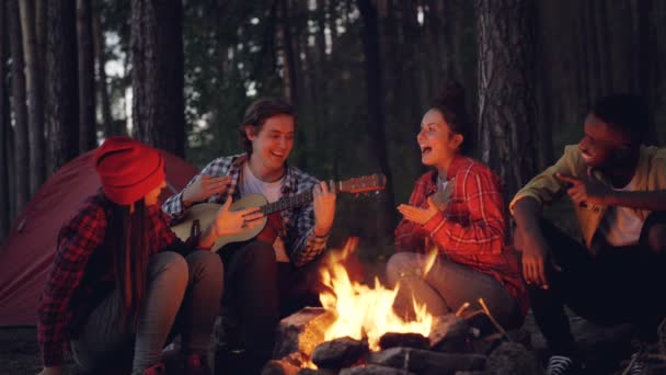 Boucle cinématographique - jeune touriste joue de la guitare pendant que ses amis chantent et rient assis autour du feu dans le bois le soir en profitant de la nature et de la compagnie . — Video