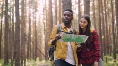 Mutlu çok ırklı çift harita, söz ve güneşli yaz aylarında vahşi ormanda kaçış sırasında gülerek bakıyor. Ağaçlar, otlar ve sırt çantaları görülebilir.