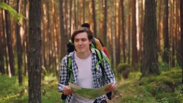 Un joven sonriente está mirando el mapa y buscando el camino correcto en el bosque mientras un grupo multiétnico de amigos lo sigue. Concepto de amistad, naturaleza y personas . — Vídeos de Stock