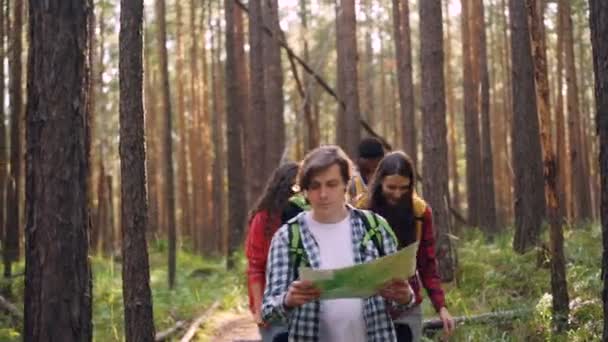Turisti felici sorridenti giovani camminano nella foresta con cartina cartacea chiacchierando e ridendo godendo della libertà e della bella natura e portando zaini . — Video Stock