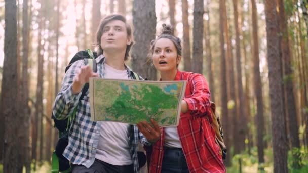 Casal de turistas mulher jovem e homem estão estudando mapa e olhando em torno de pé na floresta no dia de verão vestindo roupas casuais. Caminhadas e conceito de navegação . — Vídeo de Stock