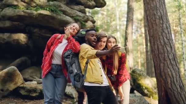 Multiracial group of friends tourists are taking selfie in forest with rocks in background using smartphone, men and women are posing and showing hand gestures. — Stock Video