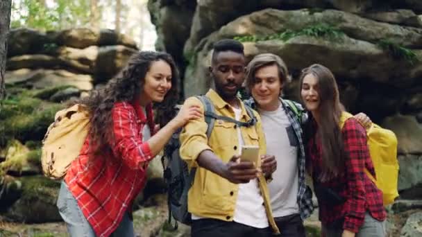 Chico afroamericano feliz está haciendo videollamadas usando un teléfono inteligente durante la caminata en el bosque con amigos, los jóvenes están mirando la pantalla, saludando con la mano y hablando . — Vídeos de Stock