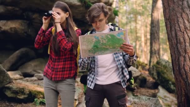 Guapo joven está mirando el mapa mientras su amigo está mirando en prismáticos, señalando un lugar interesante y sonriendo. Concepto de personas, naturaleza y juventud . — Vídeos de Stock