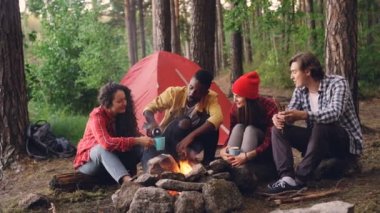 Afro-Amerikan yakışıklı Termos şişe sıcak içecek gözlük ve kamp ateşi etrafında oturan arkadaşlar çok ırklı grubuyla bardak içine dökme.