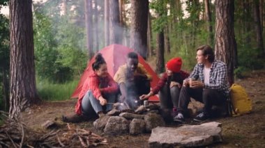 Genç erkek ve kadın gezginlerin çok ırklı grup ormanda oturan ve gülümseyerek ateşe hatmi kavurma yiyecek yemek pişiriyor. Hiking, sağlıklı yaşam tarzı ve gençlik kavramı.