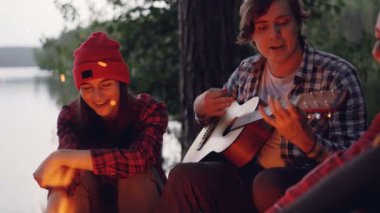 Turist çok ırklı grup ormandaki göl yakınındaki ormanda gitar ve şarkı kamp ateşi etrafında oynarken eğleniyor. İnsanlar rahat giysiler giyiyor.